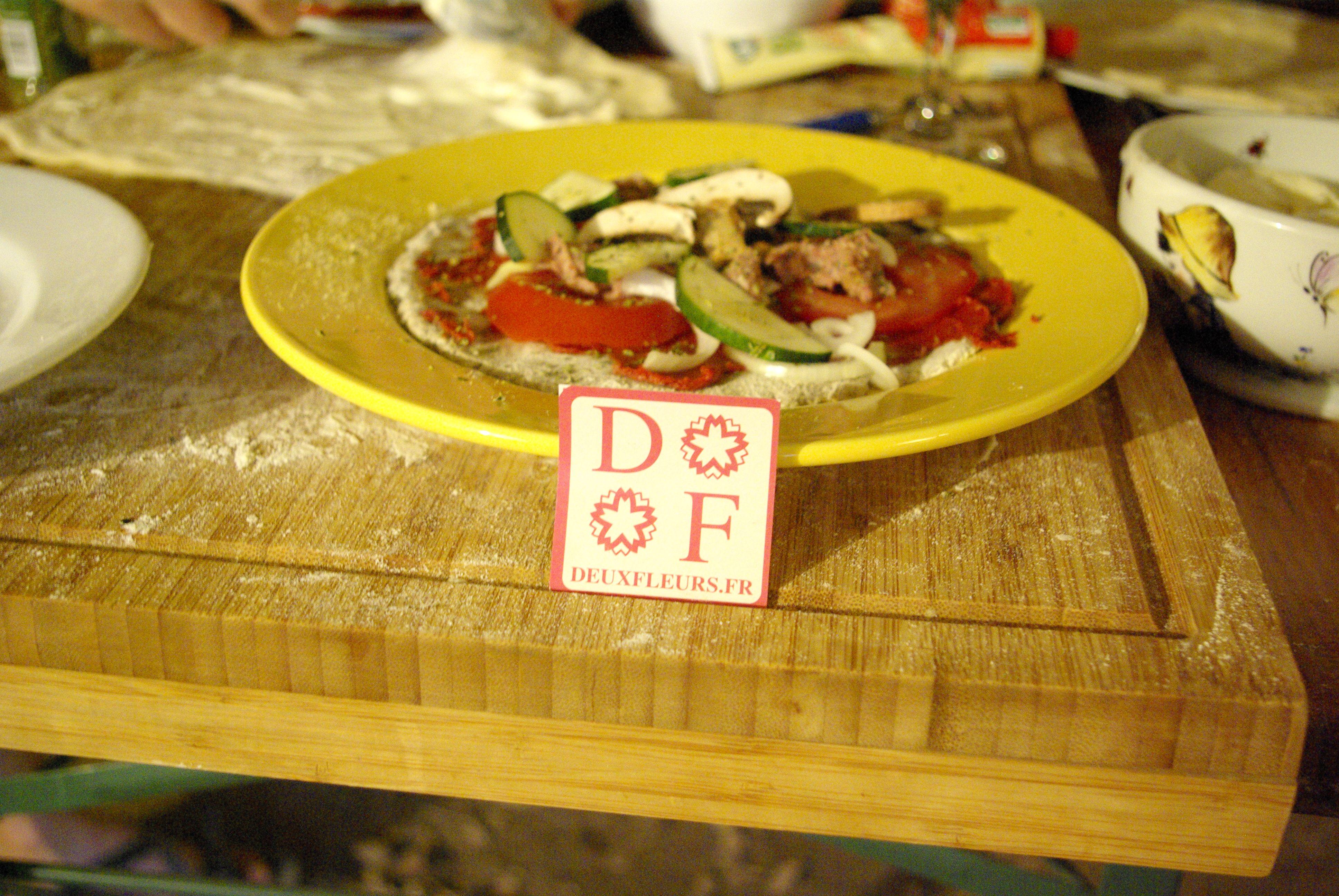 Photographie d'une petite pizza en cours de préparation. Un autocollant Deuxfleurs est posé devant, face caméra.
